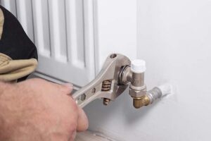 A homeowner using a wrench to replace a radiator valve on a white wall radiator during a DIY home heating fix.